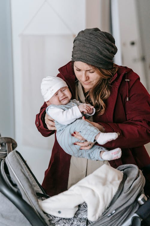 Young woman in warm clothes and hat hugging and putting cute crying newborn into stroller before going to walk