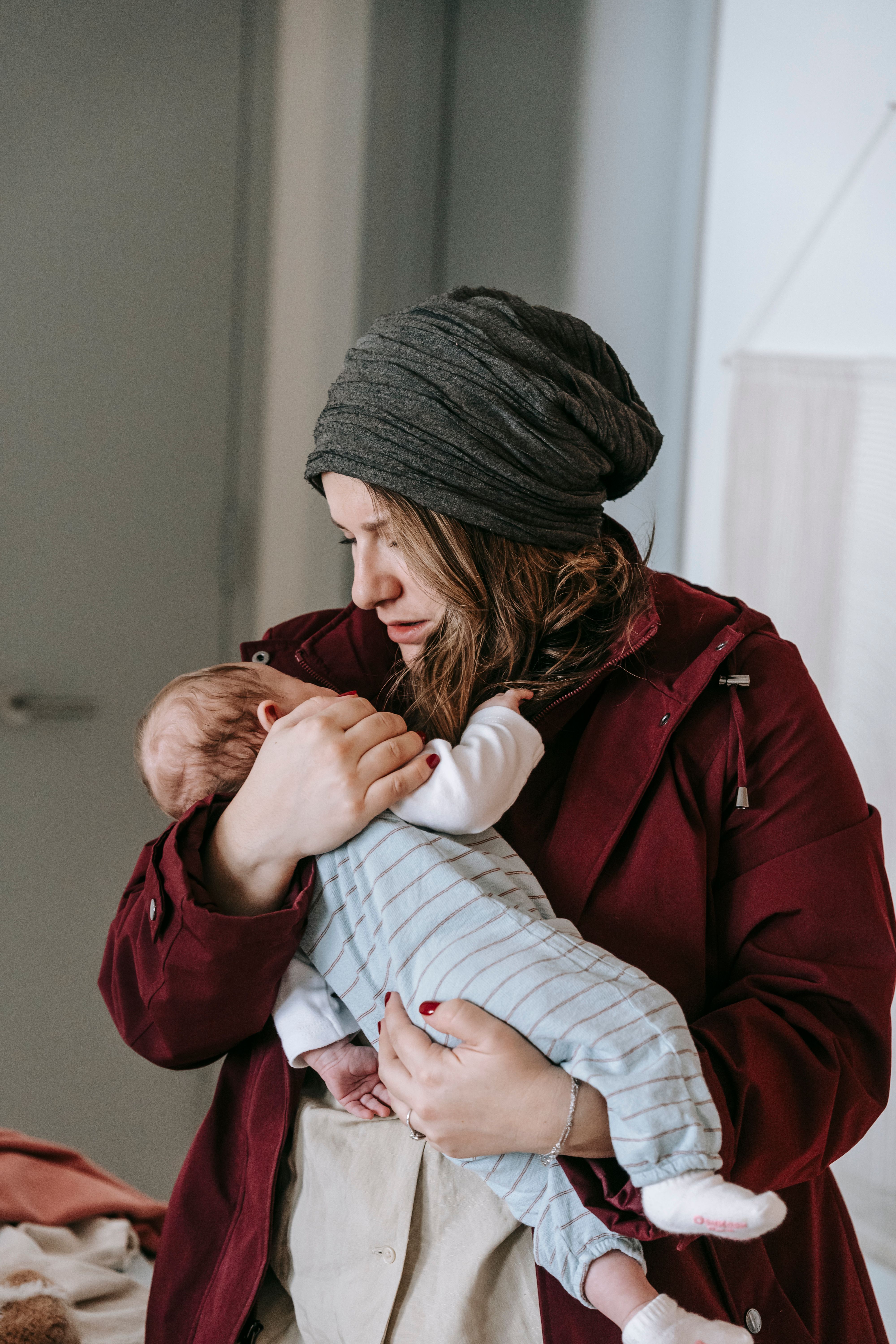 Young woman in warm clothes and hat embracing and calming adorable baby at home