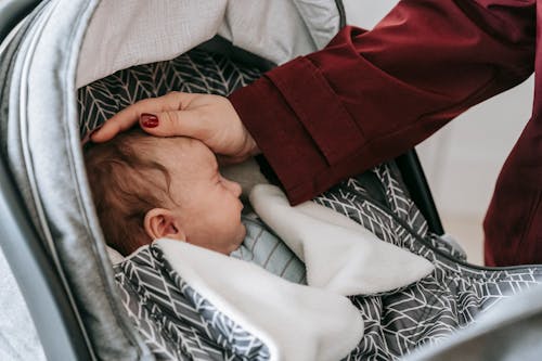 From above of crop unrecognizable female in coat caressing cute newborn sleeping in baby stroller during walk in daytime