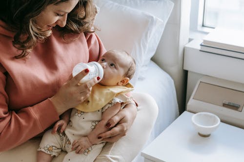 Free From above of crop happy young mother in casual clothes smiling while hugging and feeding adorable newborn from bottle sitting on comfortable bed Stock Photo