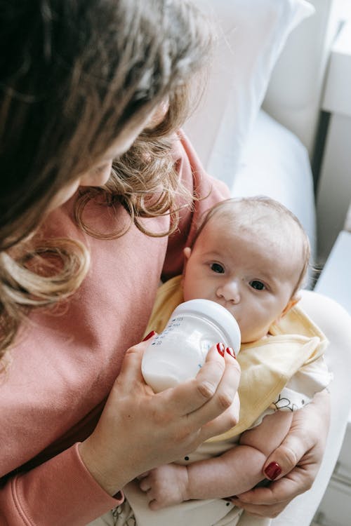 High angle of crop unrecognizable young mother in casual clothes sitting on bed and feeding adorable baby with milk from bottle in sunny morning