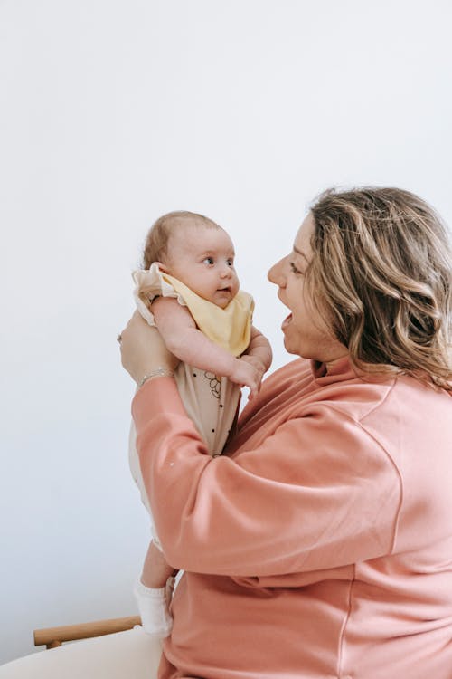 Side view of cheerful young mother in casual clothes hugging and having fun with adorable newborn in daytime at home