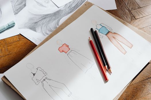 Artistic fashion design sketches on paper accompanied by colored pencils on a wooden surface.
