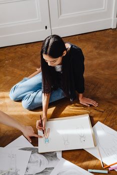 A young woman sits on the floor sketching fashion designs with pencil and paper.
