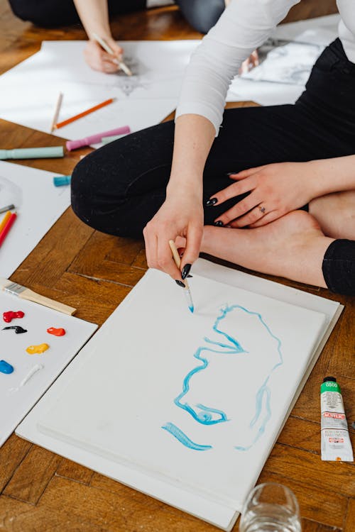 A Person Sitting on a Wooden Floor while Painting on White Paper