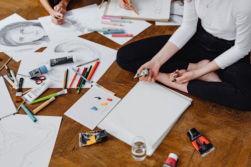 A Person in White Long Sleeves and Black Pants Sitting on a Wooden Floor Near the Scattered Art Materials