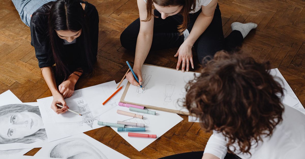 Trois femmes participent à une session de dessin collaborative sur un sol en bois, entourées de croquis et de fournitures artistiques.