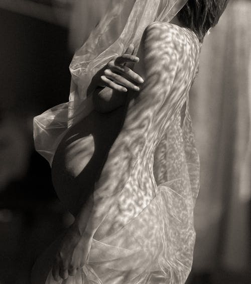 Free Black and white side view of naked anonymous pregnant female wearing transparent cloth covering breasts while standing in room with sunlight Stock Photo