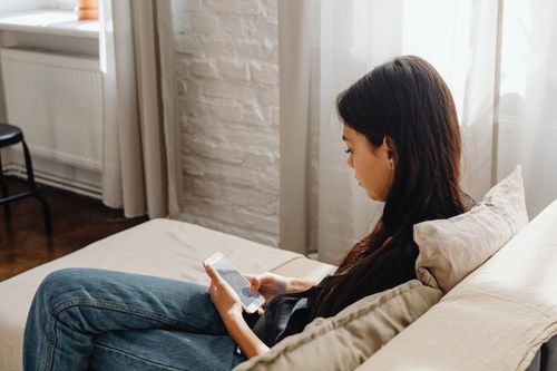Free Young woman using smartphone relaxed on sofa in cozy indoor setting. Stock Photo