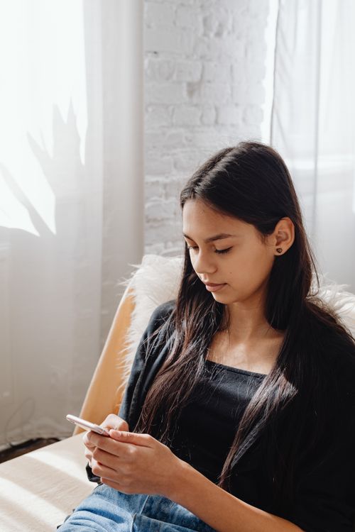 Free Woman with long hair using a smartphone indoors, sitting comfortably and focused. Stock Photo