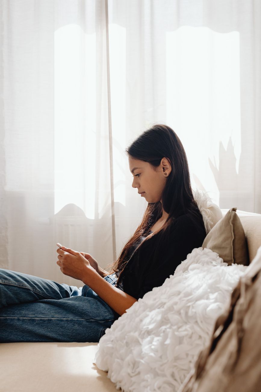 A teenage girl sits by a window checking her phone, representing the kind of online device use linked to the reported risks facing adolescents.