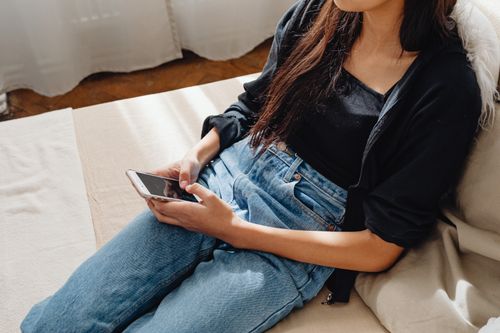 Free Young woman in casual clothing using smartphone while relaxing on a sofa indoors. Stock Photo