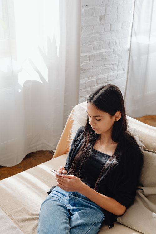 Free Young woman sitting on a sofa in a bright room, using a smartphone and enjoying leisure time. Stock Photo