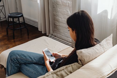Free A woman sits comfortably on a sofa, browsing her smartphone in a cozy indoor setting. Stock Photo
