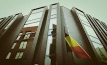 一座采用砖玻璃立面的现代化城市建筑，在多云天空下悬挂着比利时国旗