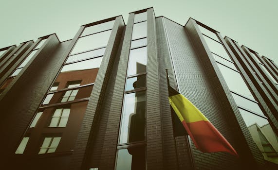 一座采用砖玻璃立面的现代化城市建筑，在多云天空下悬挂着比利时国旗