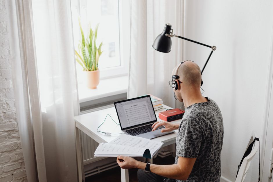Bald man working at home office with laptop, holding papers, and wearing headset.