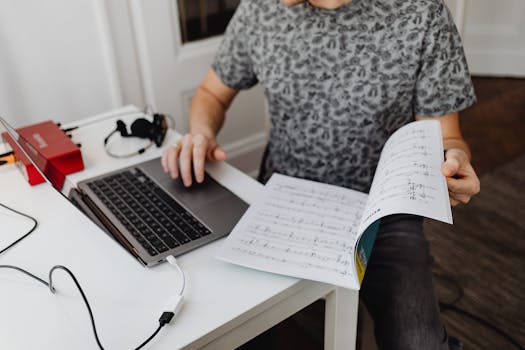 A person typing on a laptop while holding music sheets, focused on work.