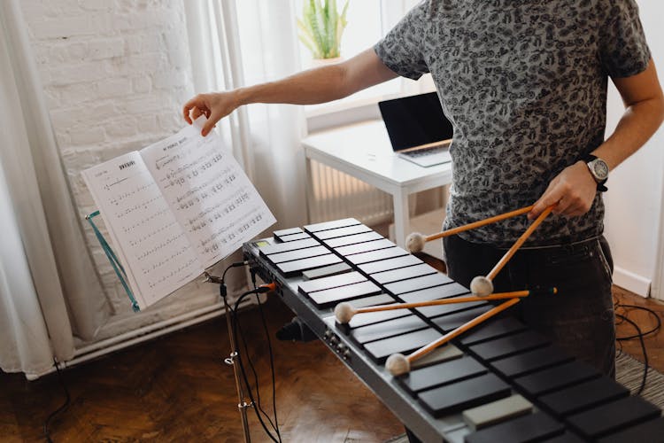 A Person Turning Pages Of A Music Book While Holding Mallets