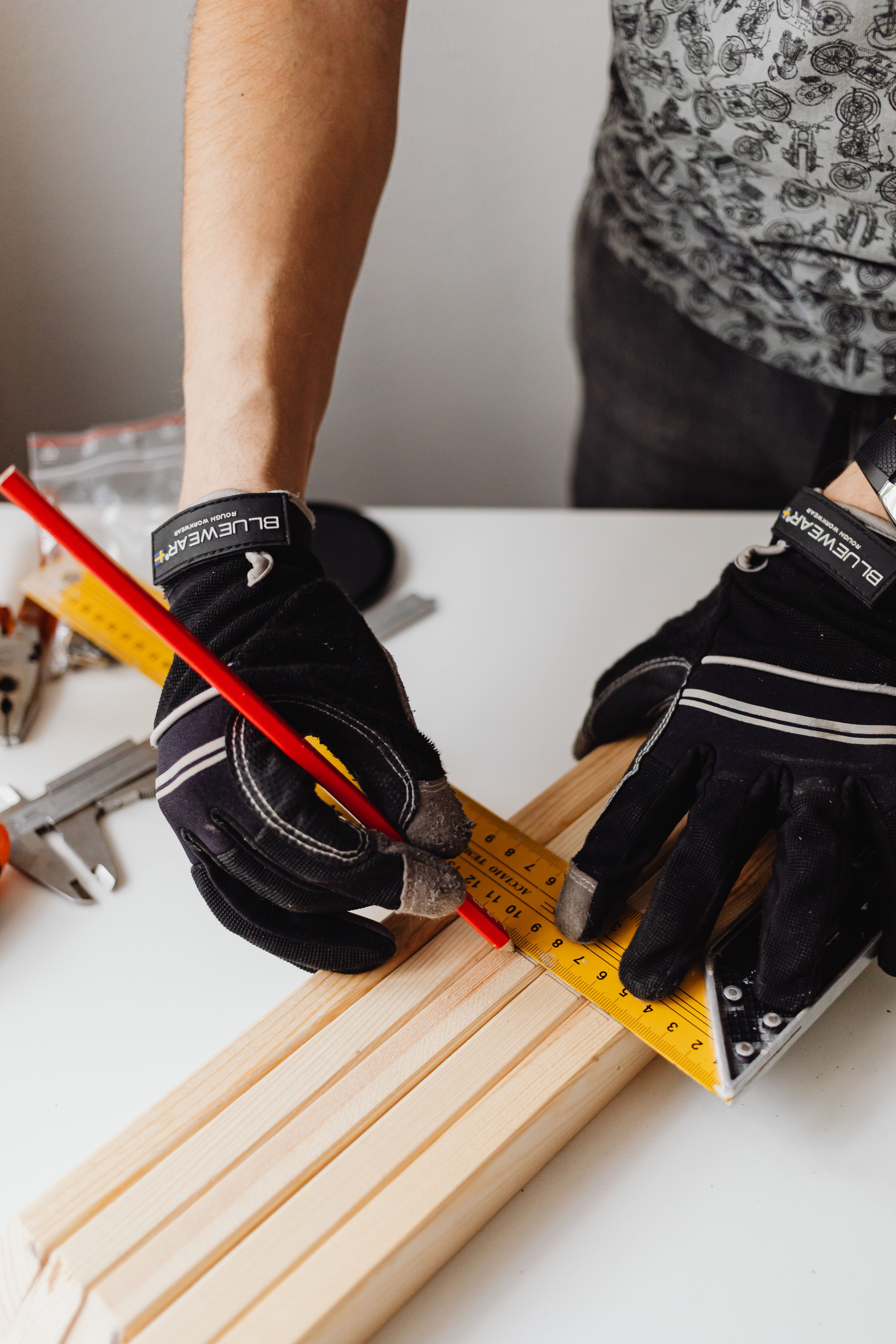 Gloved craftsperson marking and measuring a wooden plank with a square ruler and pencil