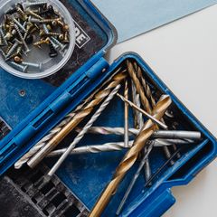 Open blue case of gold titanium and silver drill bits beside a tub of screws on a pale blue sheet
