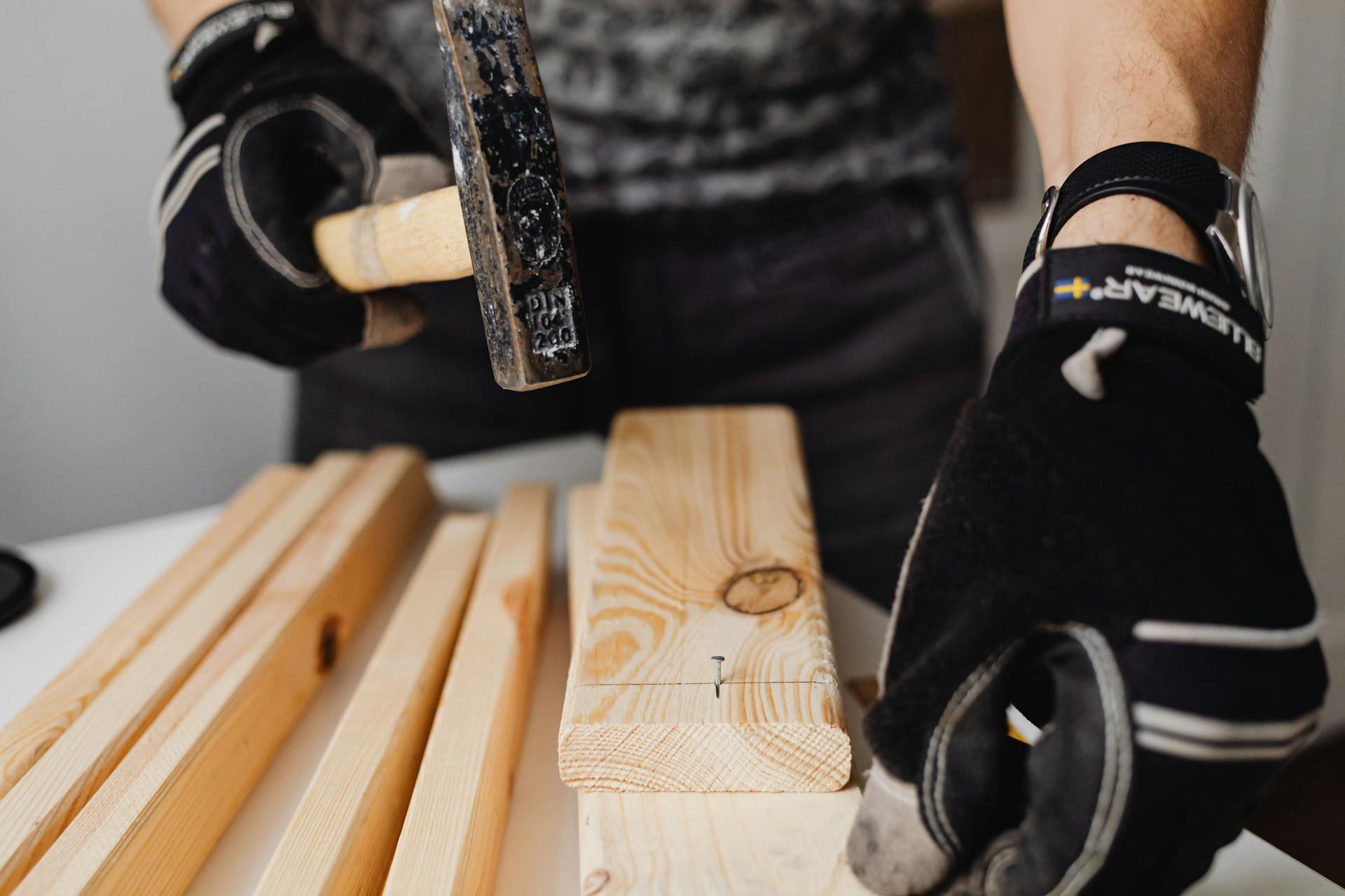 A carpenter wearing gloves uses a hammer to drive nails into wooden planks in a workshop.