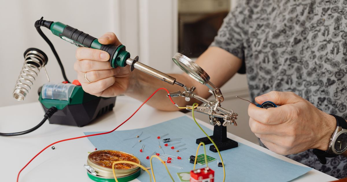 Hands soldering wires held in helping hands with a magnifier, a soldering station and coil iron stand on a bright white desk