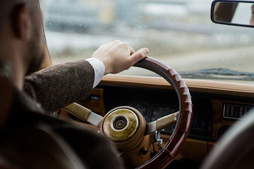 Crop faceless man in stylish clothes sitting in car on seat with hand on steering wheel in daylight while driving on street
