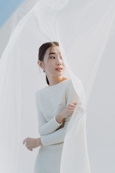 Portrait of a young woman in a white dress behind a sheer veil in a studio setting.