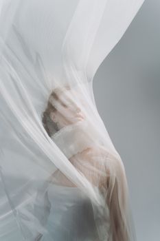 Ethereal portrait of a woman wrapped in sheer fabric on a soft gray background, creating a dreamlike effect.