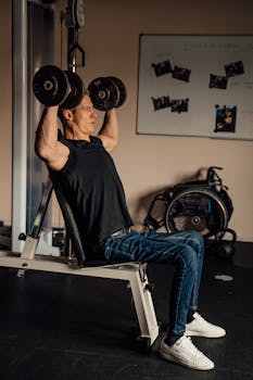 Muscular man in a gym performing shoulder press exercises with dumbbells, showcasing fitness and strength.