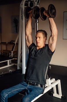 A muscular man lifting dumbbells while sitting inside a gym, focused on his workout.