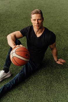 Athletic man in casual wear sitting with a basketball outdoors on grass.
