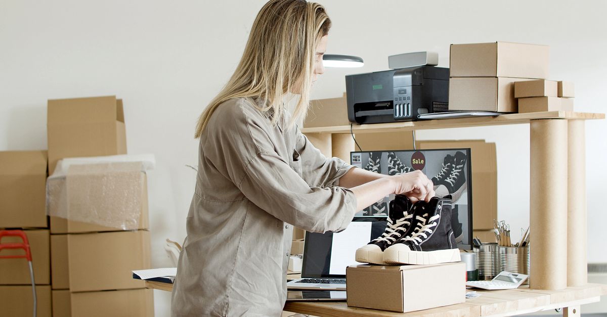 Žena balí tenisky do krabice na svém e-commerce pracovišti obklopená balicím materiálem.