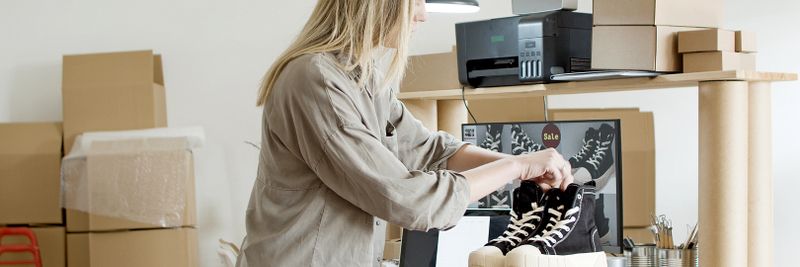 A woman packs sneakers in a box at her e-commerce workspace, surrounded by packing materials.