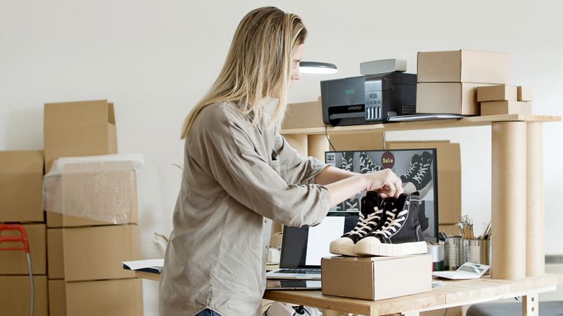 A woman packs sneakers in a box at her e-commerce workspace, surrounded by packing materials.