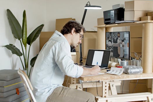 Businessman managing an online store in a home office setting, typing on a laptop with packaging supplies around.
