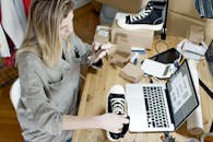 Woman photographing shoes for online sale in home workspace with laptop and packages.