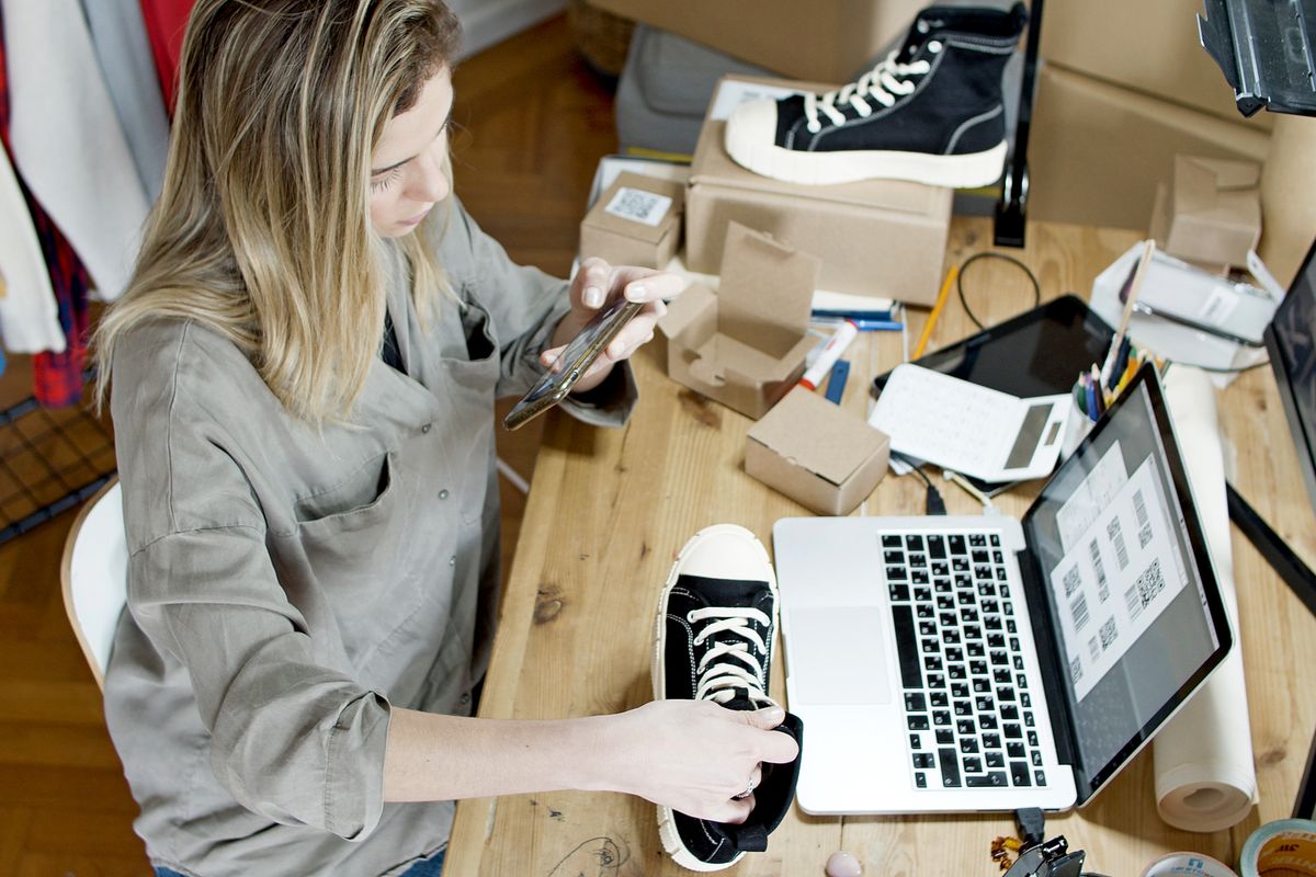 Empreendedora fotografando um tênis com o celular para anunciar em uma loja virtual, com laptop e caixas de papelão na mesa