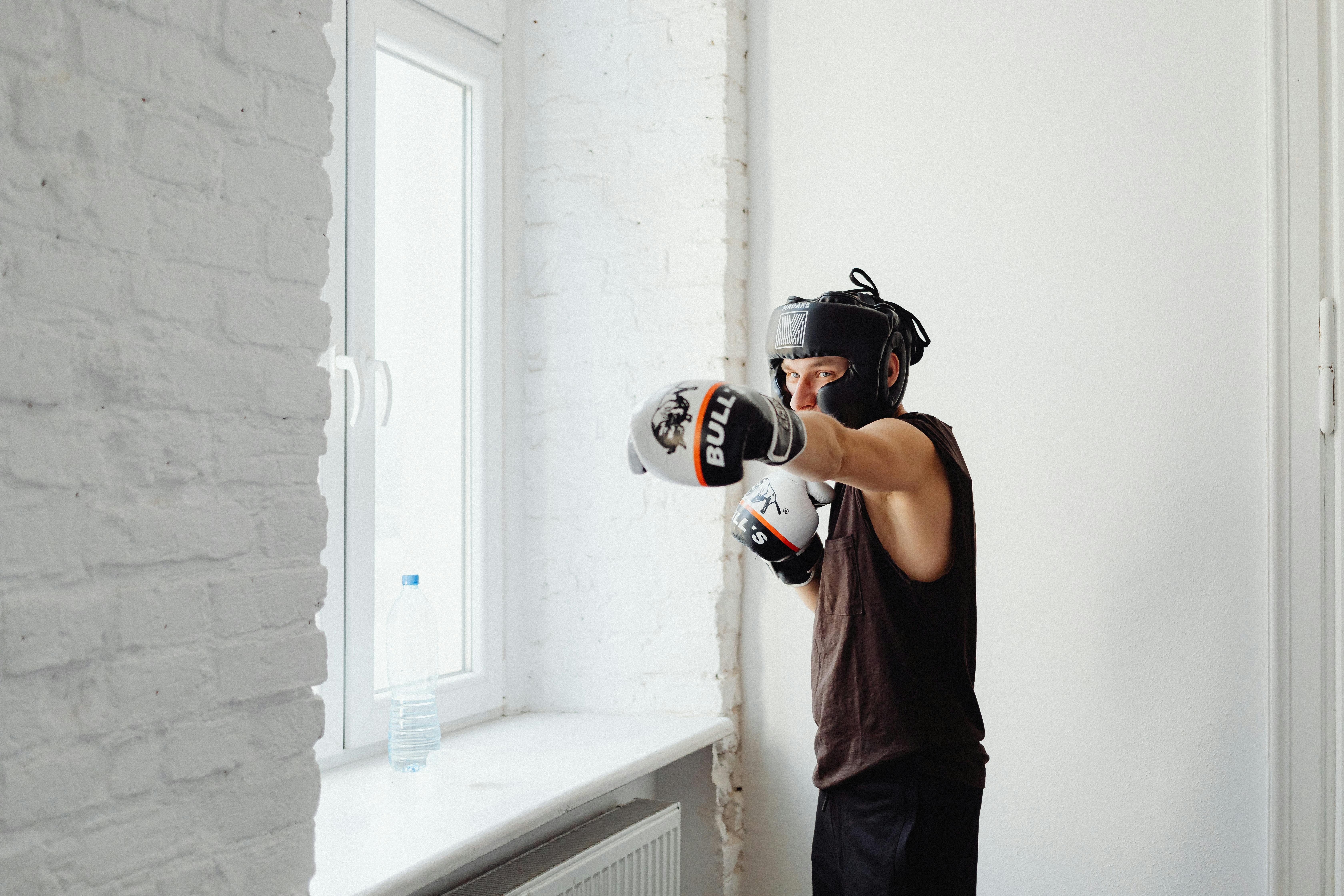 A male boxer practices punches indoors, wearing gloves and headgear for safety.