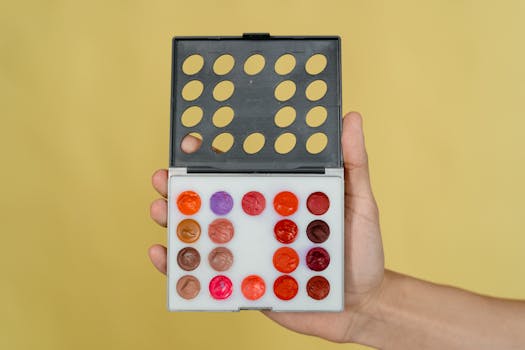 Hand holding a vibrant makeup palette with lipstick shades against a yellow backdrop.