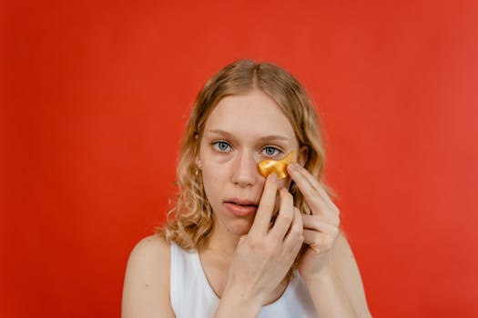Close-up portrait of a woman applying an under eye mask against a red background, focusing on skincare routine.