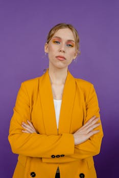 Confident woman in a yellow blazer posing with arms crossed against a purple background.