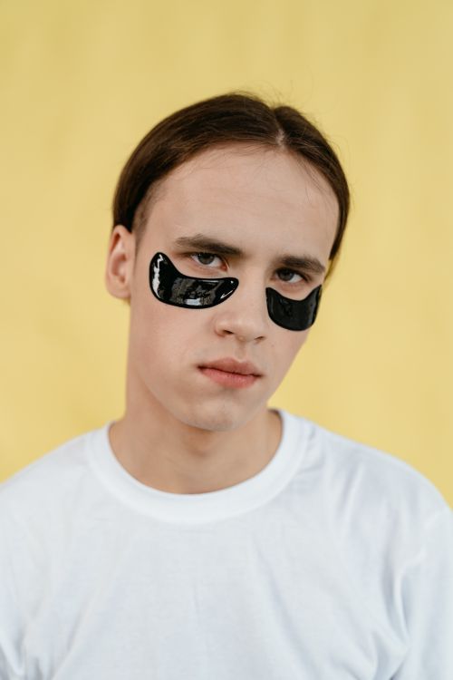 Free Young man wearing black under-eye patches against a vibrant yellow background, promoting skincare routine. Stock Photo