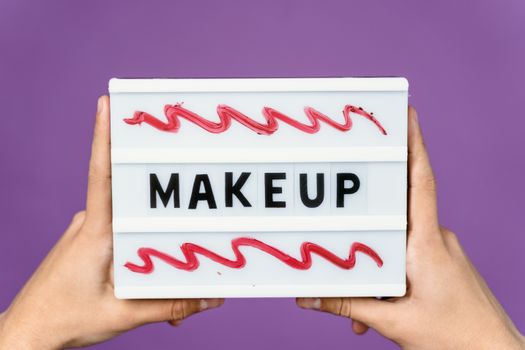 Close-up of a lightbox with the word 'MAKEUP' held by hands, featuring artistic red swirls.