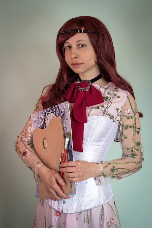 Free Young female artist in dress with floral ornament and neckerchief holding palette picture and art brushes and looking at camera Stock Photo