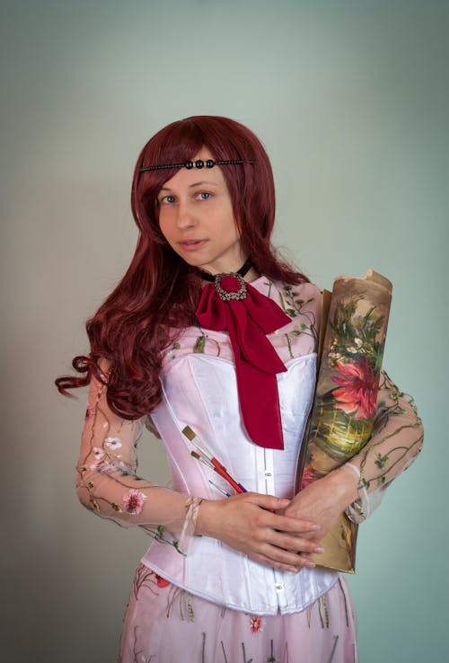 Positive female with long red hair necktie wearing floral dress with white corset carrying rolled picture in canvas and art brushed