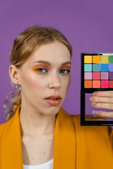 Model showcasing colorful makeup palette against a purple background in a studio setting.