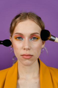 Close-up portrait showcasing vibrant eye makeup with brushes against a purple background.