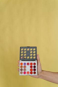 A hand displaying a vibrant makeup palette against a yellow background.